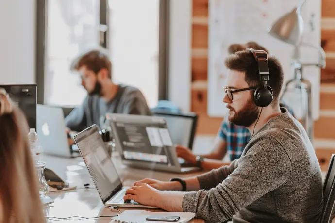 Due giovani, uno con occhiali e cuffie, lavorano su computer portatili in un ufficio con sfondo sfocato