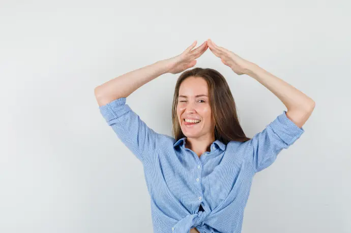 Giovane donna in camicia blu che fa il gesto del tetto della casa sopra la testa e sembra allegra